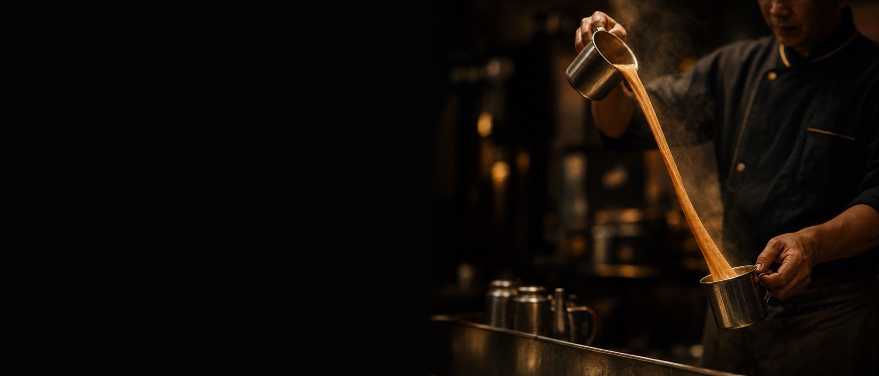 A cook pulling teh tarik between two metal cups in a dark kitchen